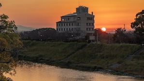 A multi-story building with a flat roof, situated beside a body of water, likely a river, with a sunset in the background. - Chikugofunagoya Kouennoyado (Chikugo)