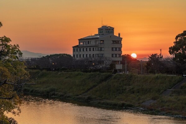 Un edificio de varios pisos con techo plano, situado junto a un cuerpo de agua, probablemente un río, con un atardecer de fondo.