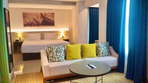 A hotel room with a bed, a sofa with patterned cushions, a small round table, and a painting on the wall. - Jambuluwuk Thamrin Hotel (Jakarta)