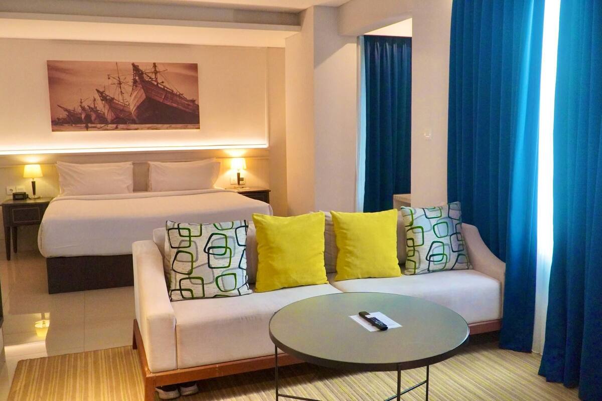 A hotel room with a bed, a sofa with patterned cushions, a small round table, and a painting on the wall.
