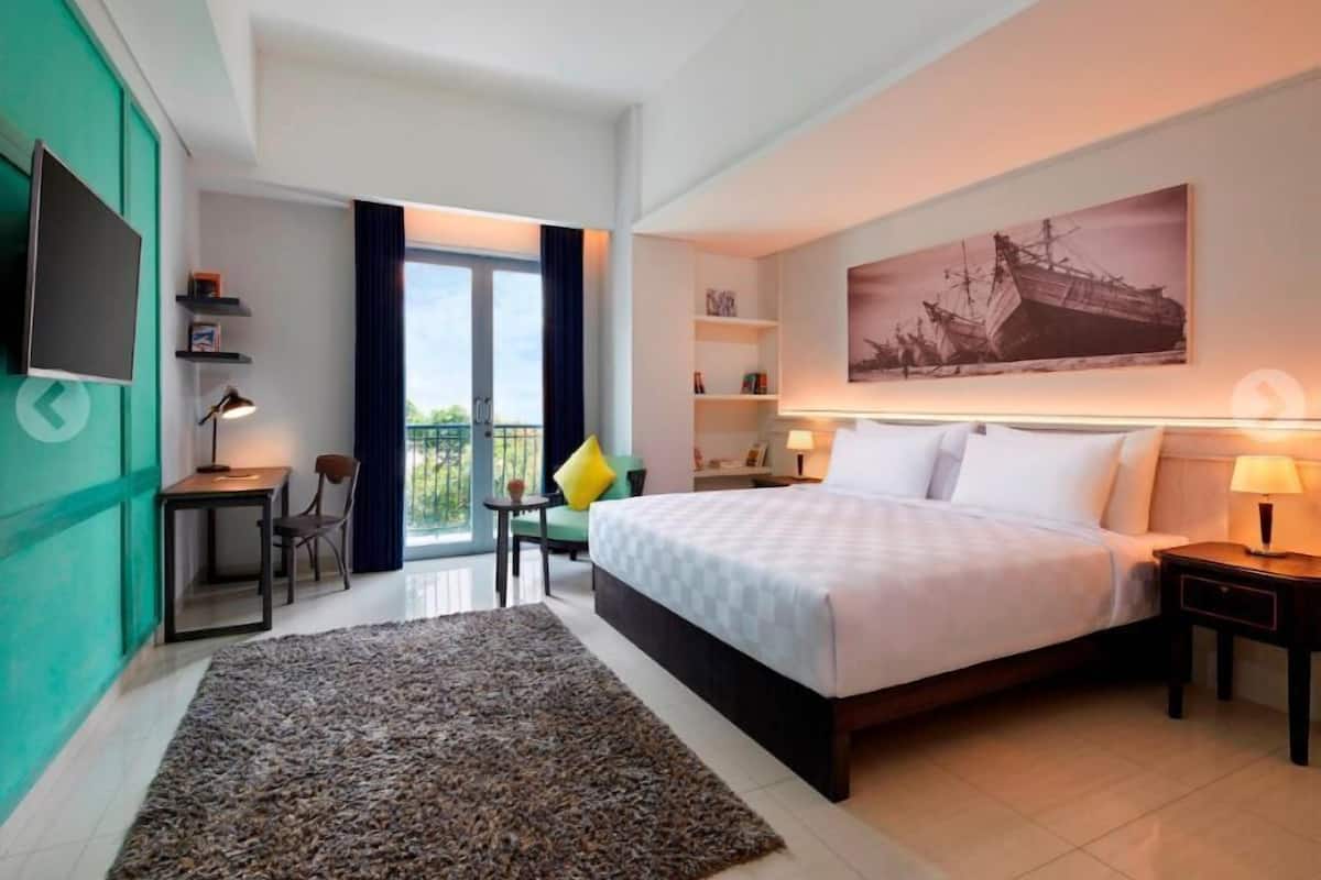 A hotel room with a bed, a desk, a chair, a TV, a bookshelf, a painting, and a balcony with curtains.