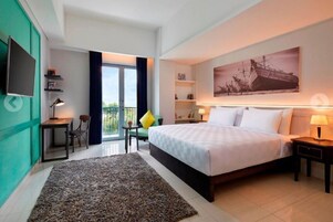 A hotel room with a bed, a desk, a chair, a TV, a bookshelf, a painting, and a balcony with curtains.