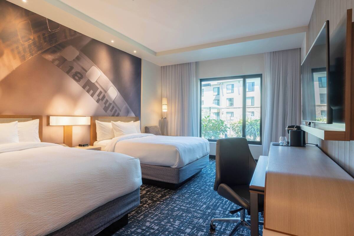 A hotel room with two beds, a desk, a chair, a TV, and a large window with a view of buildings.