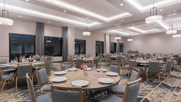 Uma grande sala de banquetes com mesas redondas preparadas para refeições, com cadeiras estofadas cinza e mesas de madeira.