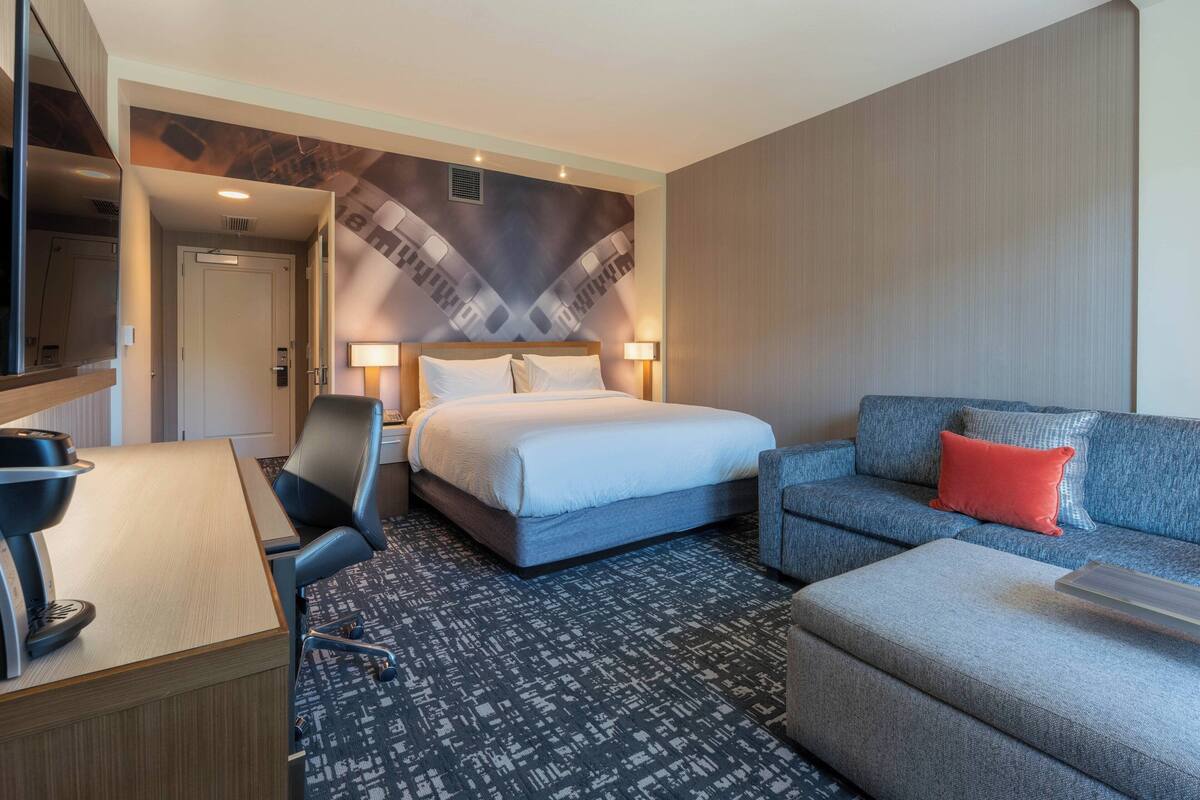 A hotel room with a bed, a desk, a chair, a sofa, and a patterned carpet.