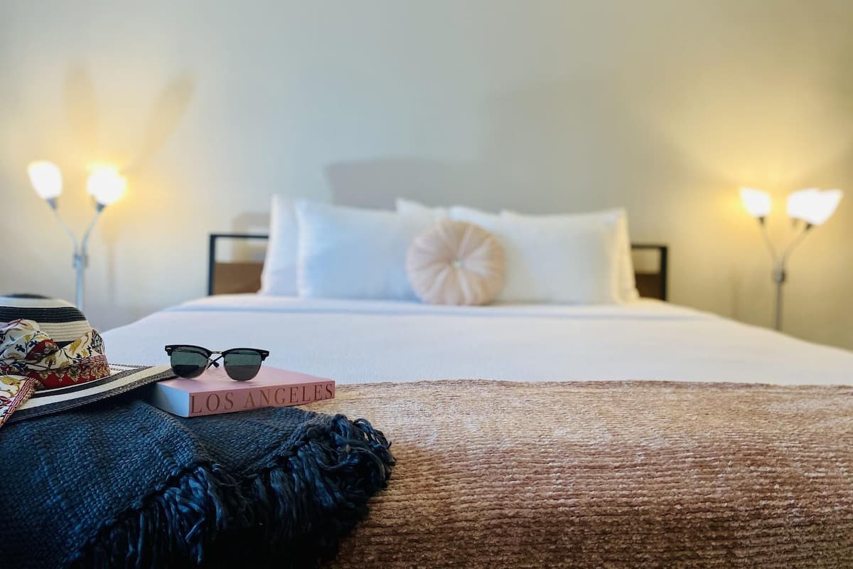 A neatly made bed with a white comforter and pillows, a book titled "Los Angeles" on the side, and a pair of sunglasses.