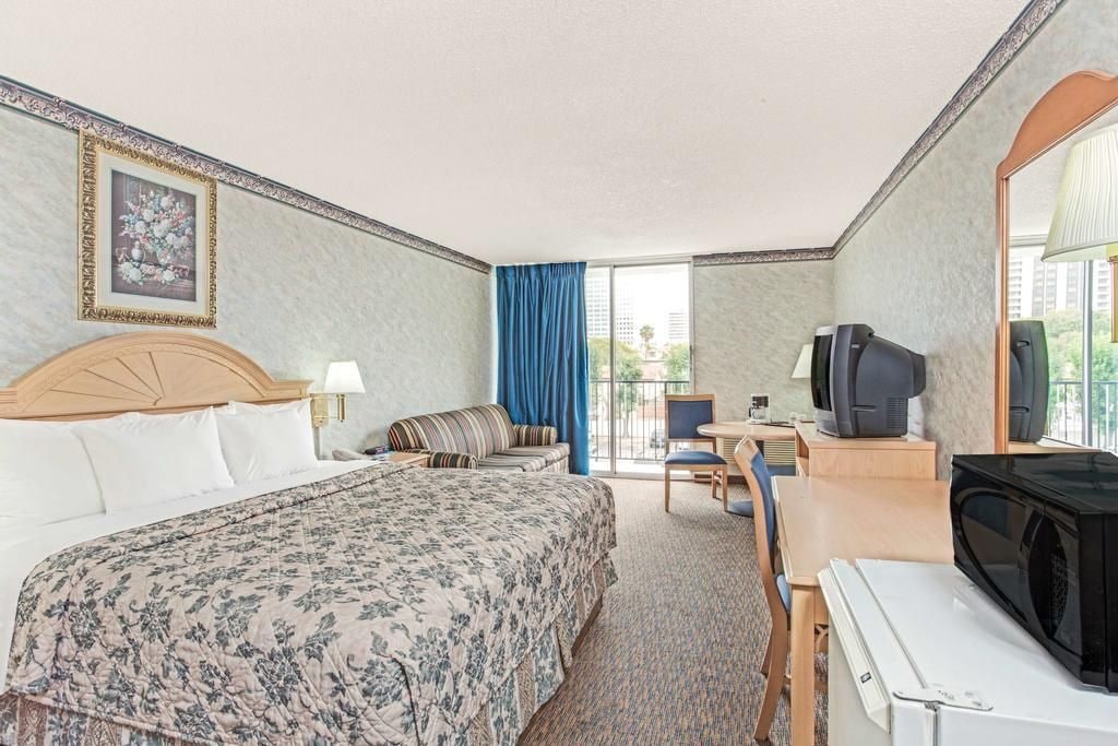 Ein Hotelzimmer mit einem Bett, einem Schreibtisch mit Fernseher, einem Sessel und einem Fenster mit Vorhängen.