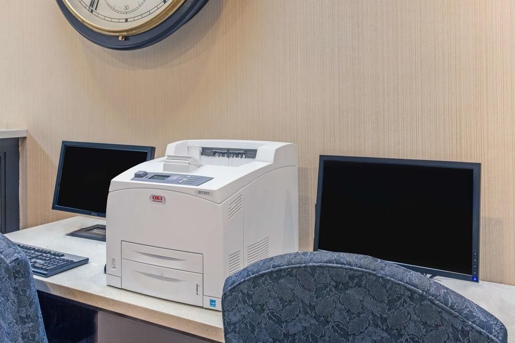 Ein Schreibtisch mit einem Drucker, zwei Computermonitoren und einer Uhr an der Wand.