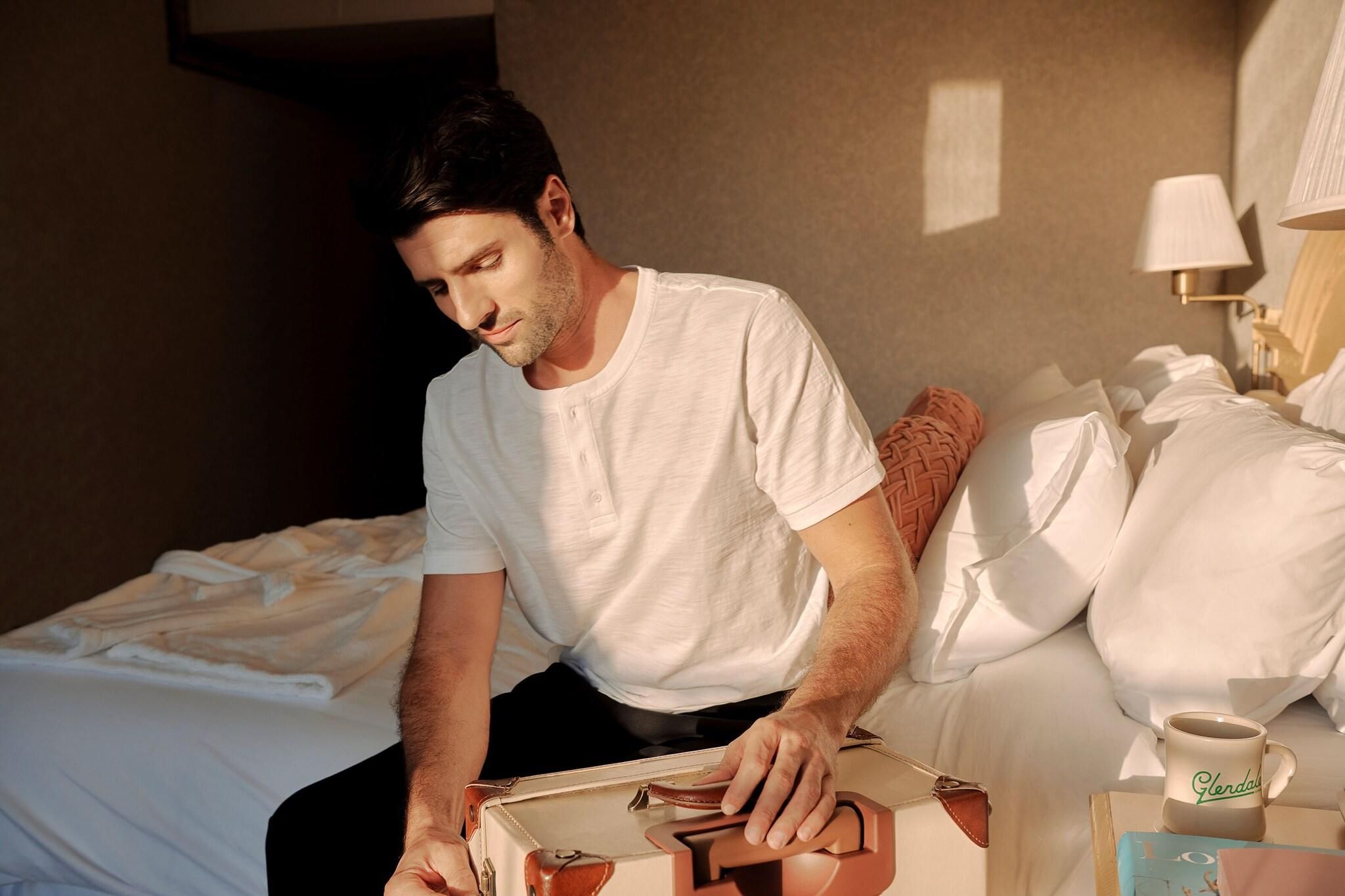 A man sitting on a bed in a hotel room, with a coffee mug and a book on the bedside table.