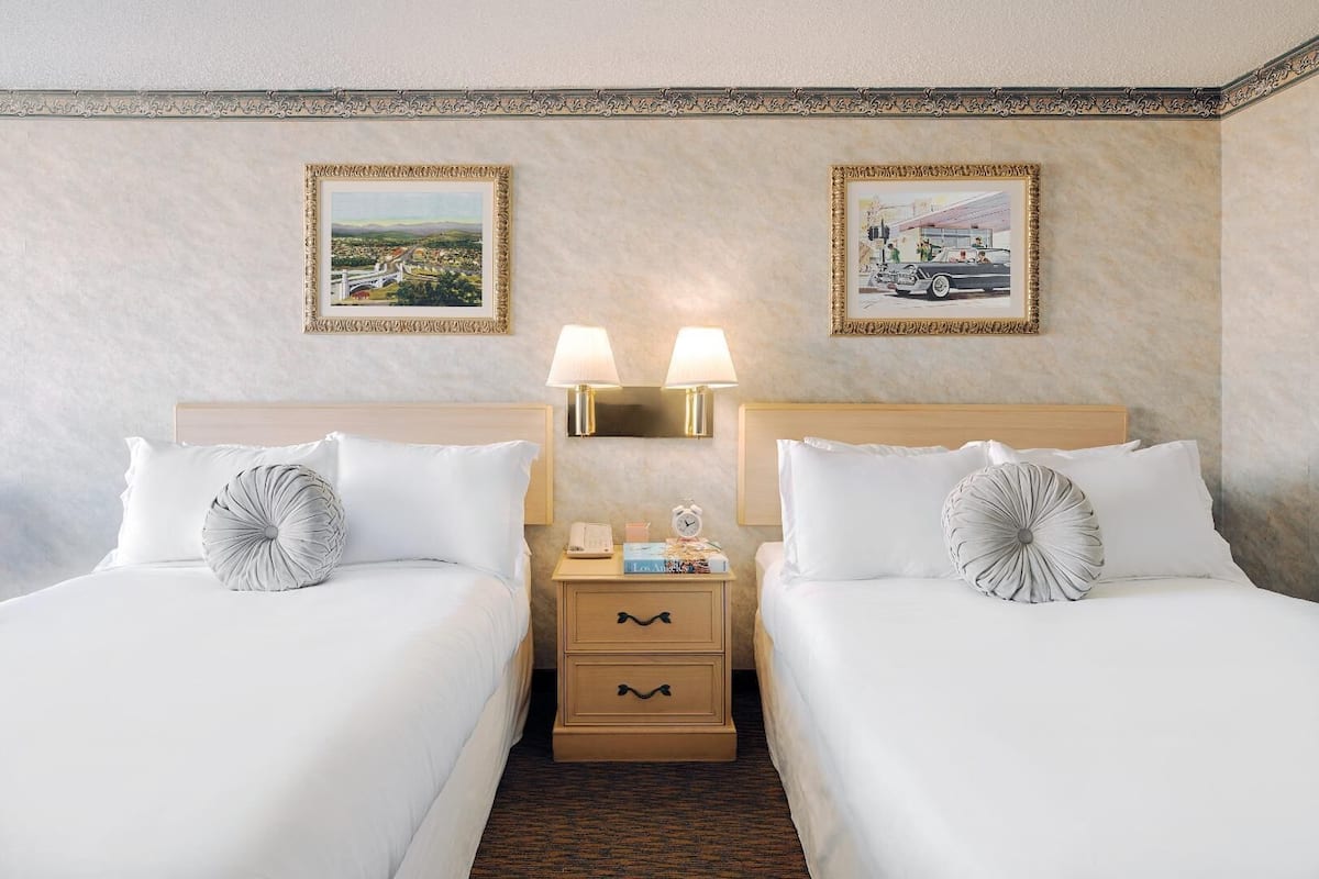 A hotel room with two beds, a nightstand with a lamp, a small table with a clock, and two framed pictures on the wall.