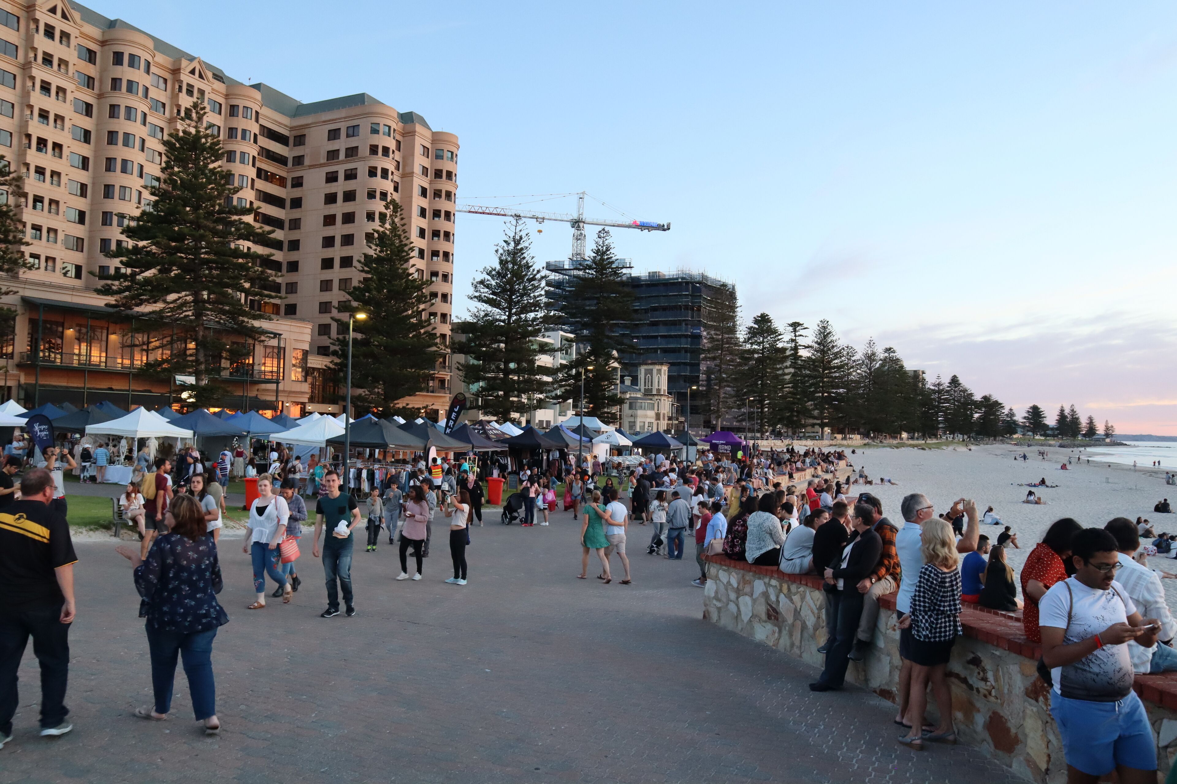 A busy waterfront promenade with people walking and socializing, a construction crane, and a beach in the background.
