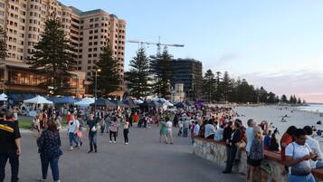 A busy waterfront promenade with people walking and socializing, a construction crane, and a beach in the background.