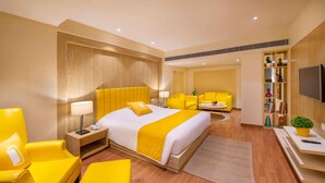 Habitación de hotel con una cama grande, sillones amarillos, un sofá, un televisor y una estantería.