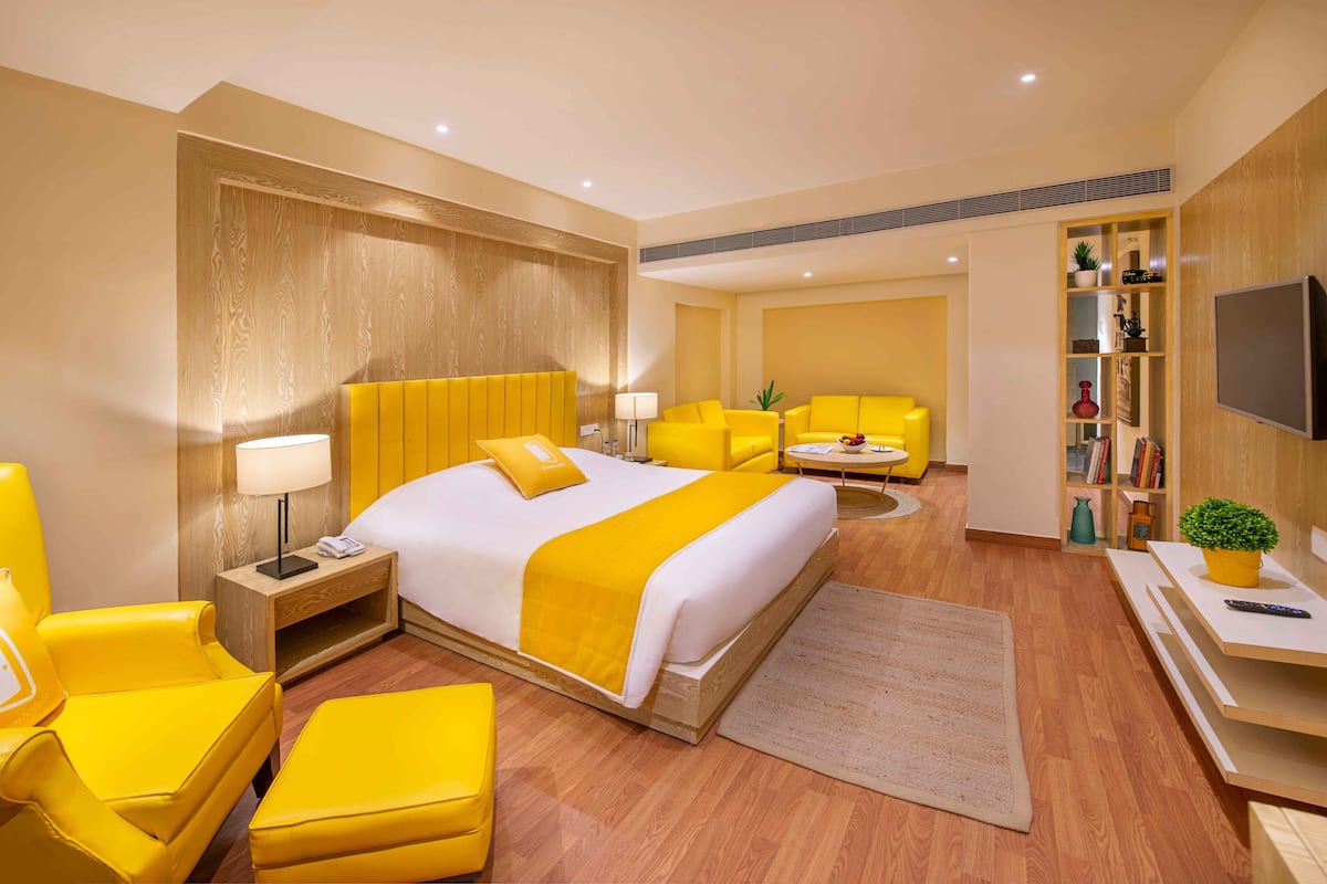 Habitación de hotel con una cama grande, sillones amarillos, un sofá, un televisor y una estantería.