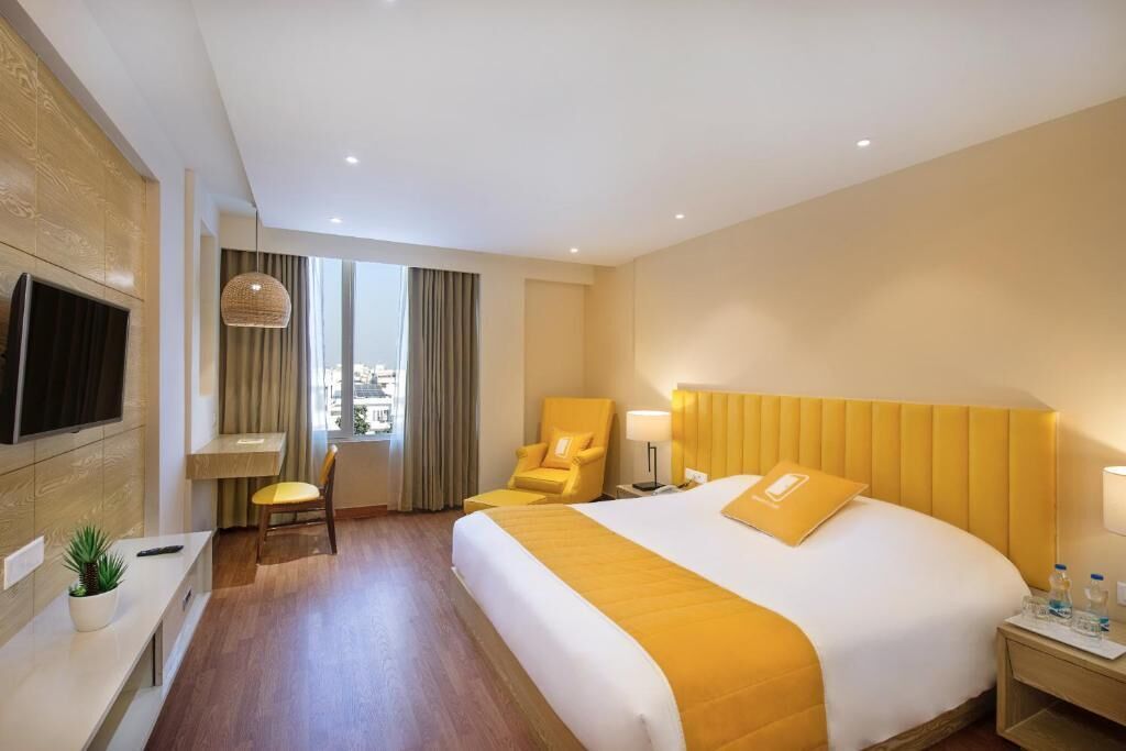 Quarto de hotel com cama, escrivaninha, cadeira, televisão e janela com cortinas.