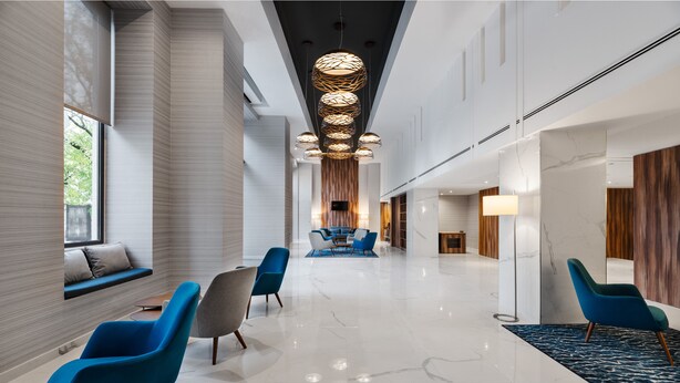 Un moderno vestíbulo de hotel con un largo pasillo, sillas azules y grises, una zona de estar central y un candelabro.