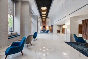 A modern hotel lobby with a long corridor, blue and grey chairs, a central seating area, and a chandelier.