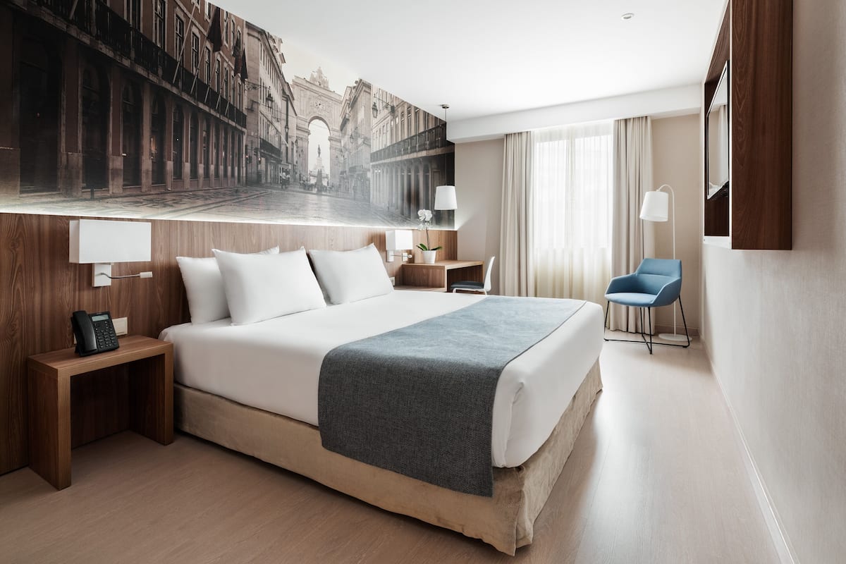 Habitación de hotel con una cama grande, una mesita de noche con teléfono, una silla, un escritorio y un ventanal con cortinas.