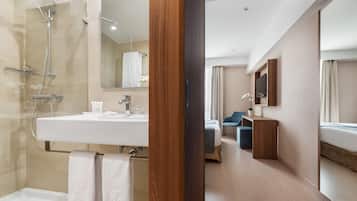 Un baño moderno con ducha, lavamanos e inodoro. Una puerta de madera da paso a un dormitorio con cama y televisor.