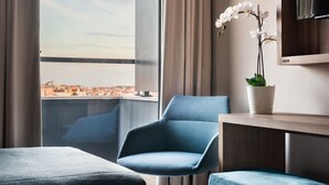 Una habitación de hotel moderna con un amplio ventanal que ofrece vistas a la ciudad, un sillón azul, una mesita con una planta en maceta y un televisor de pantalla plana montado en la pared.