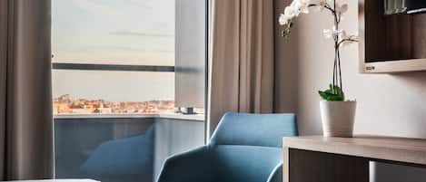 Quarto de hotel moderno com uma grande janela que oferece vista da cidade, uma poltrona azul, uma pequena mesa com uma planta em vaso e uma televisão de tela plana fixada na parede.