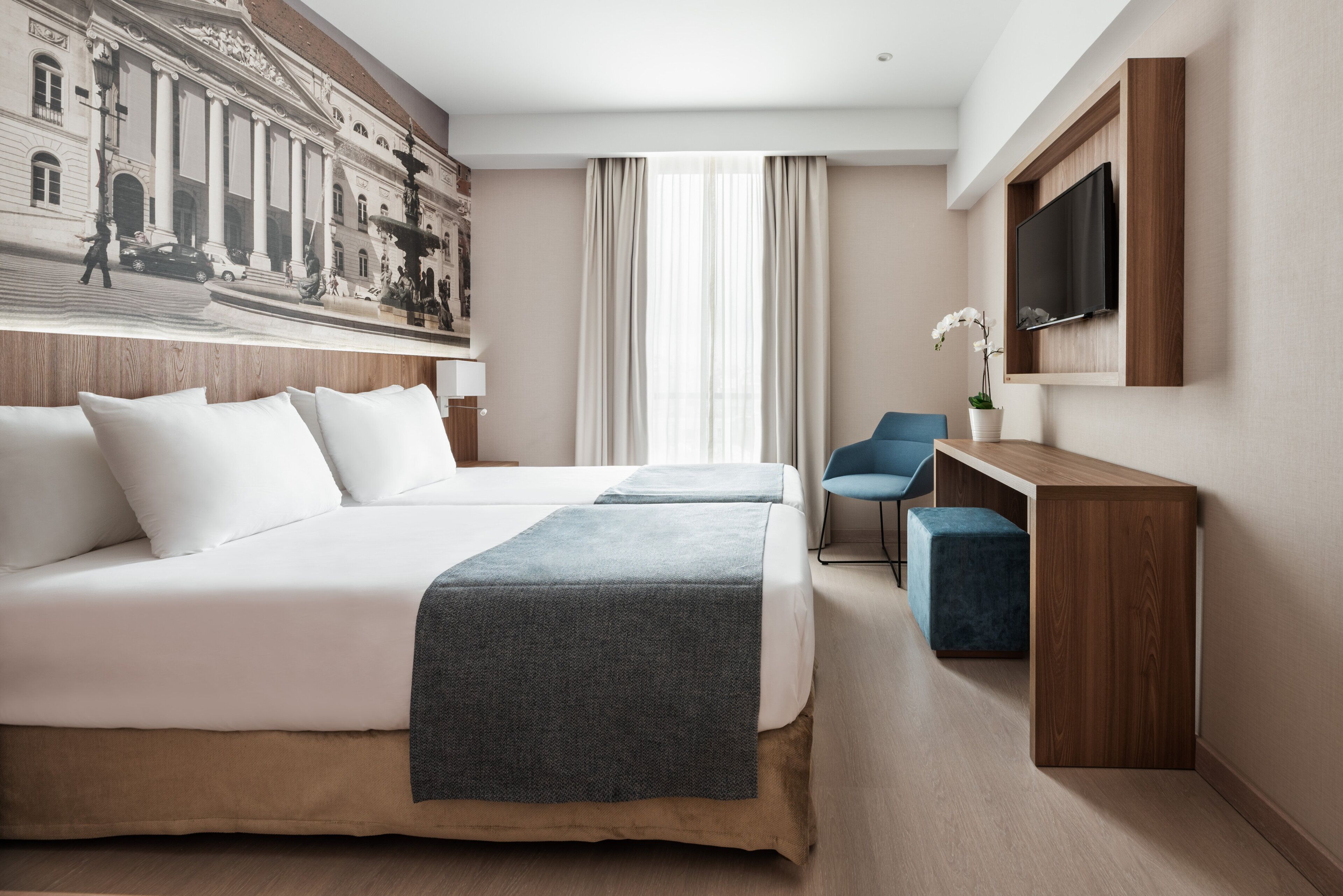 Quarto de hotel com duas camas, uma escrivaninha, uma cadeira, uma televisão e uma janela grande com cortinas.