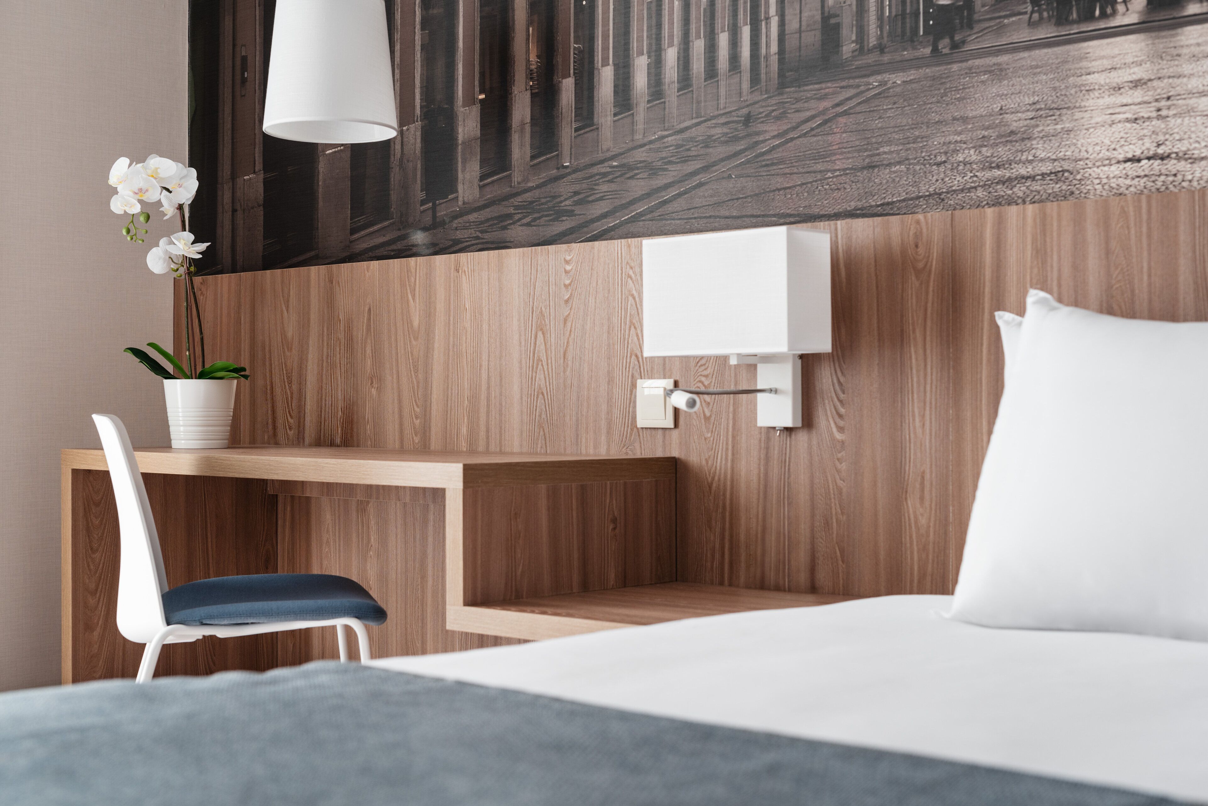 Une chambre d’hôtel avec un bureau en bois, une chaise blanche, un lit avec des oreillers blancs et une lampe fixée au mur.