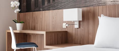 Habitación de hotel con escritorio de madera, una silla blanca, cama con almohadillas blancas y una lámpara fijada a la pared.