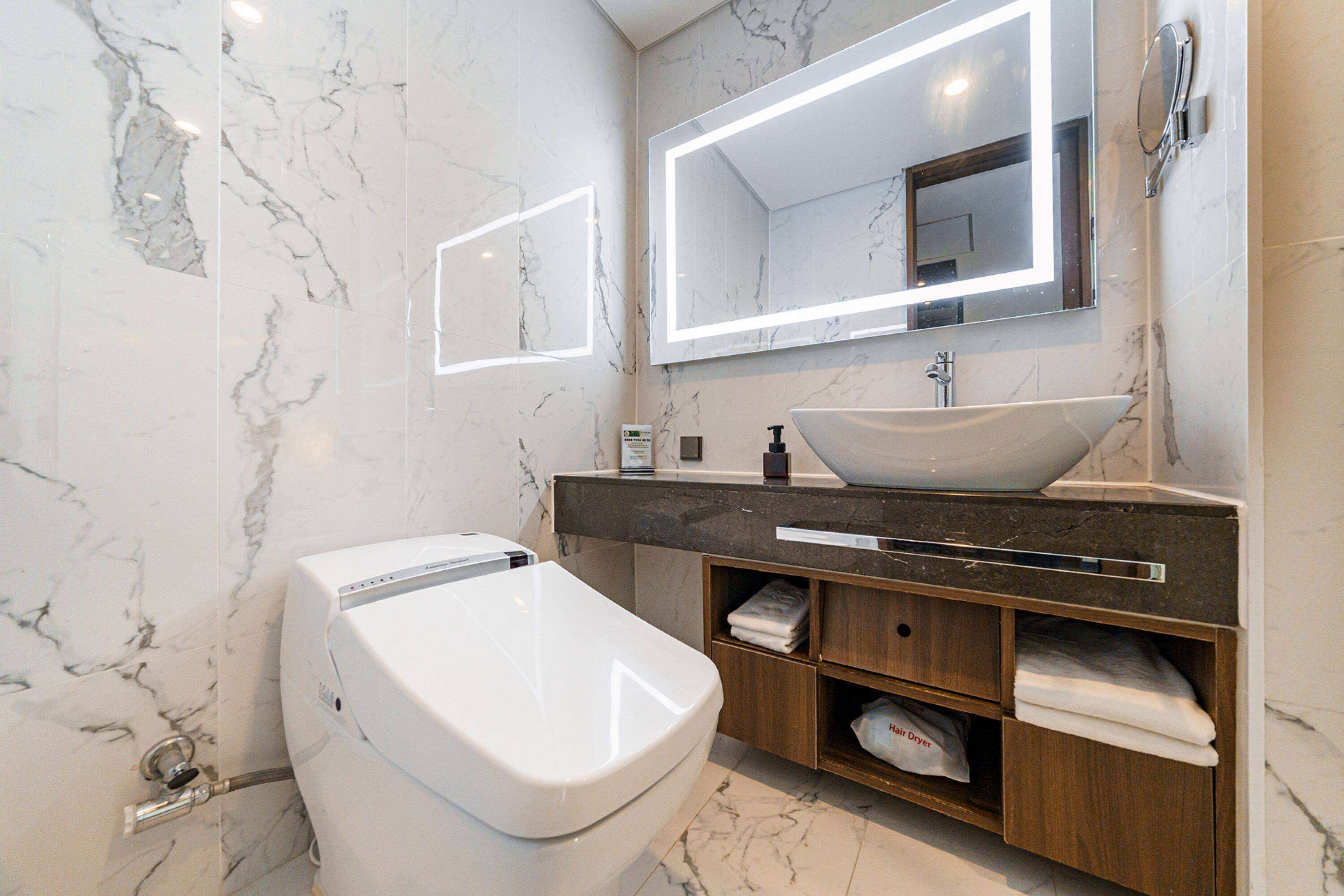A modern bathroom with a marble floor, a white toilet, a vanity with a sink, and a large mirror with a light strip.