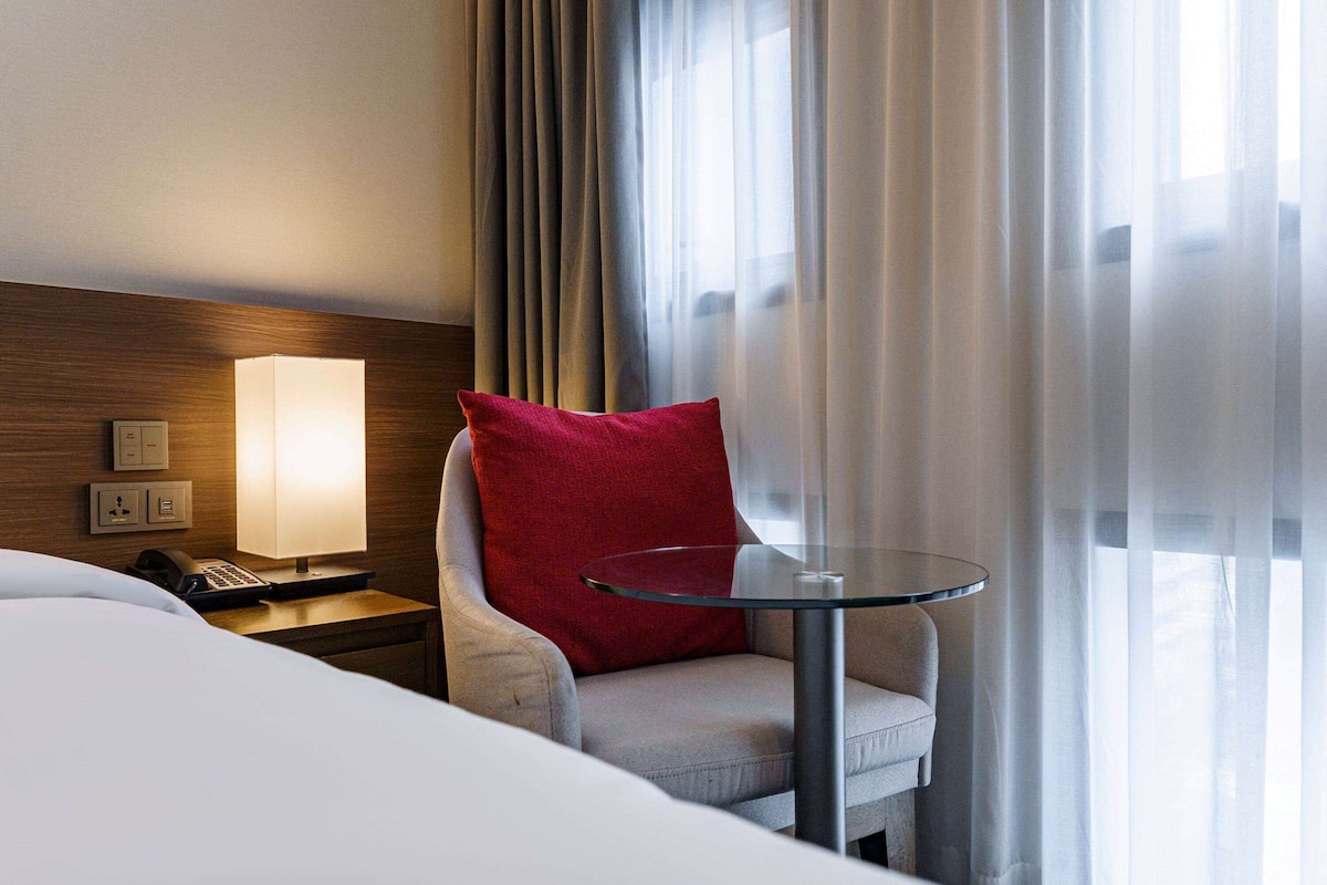 Una habitación de hotel con una cama, una silla con un cojín rojo, una mesita de noche con una lámpara y una mesa de vidrio.