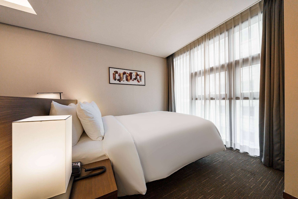 Una habitación de hotel con cama, mesita de noche, lámpara y ventana con cortinas.