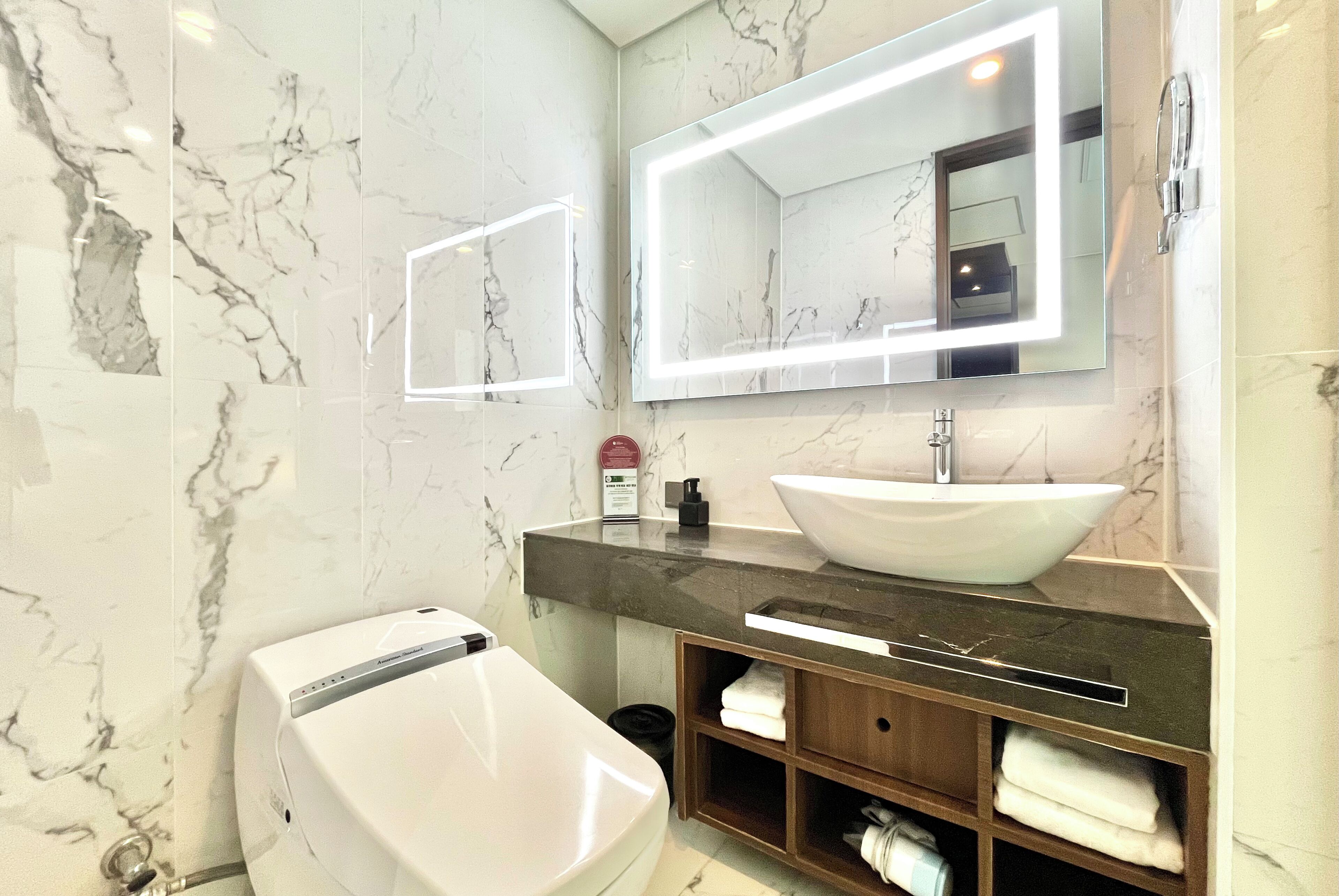 A modern bathroom with a white toilet, a large mirror with integrated lighting, a white sink, and a marble-tiled wall.