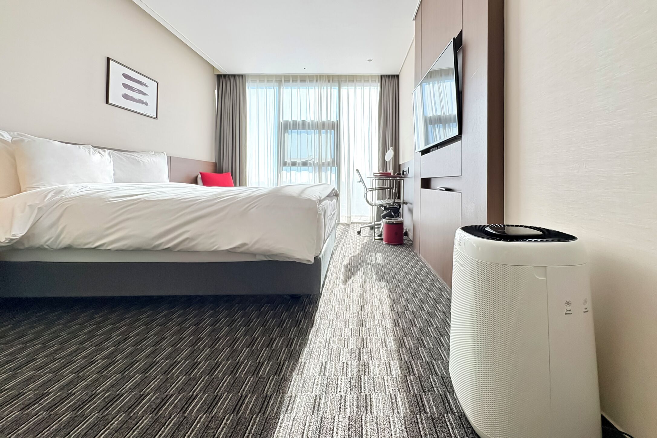 A hotel room with a large bed, a TV mounted on the wall, a standing heater, and a window with curtains.