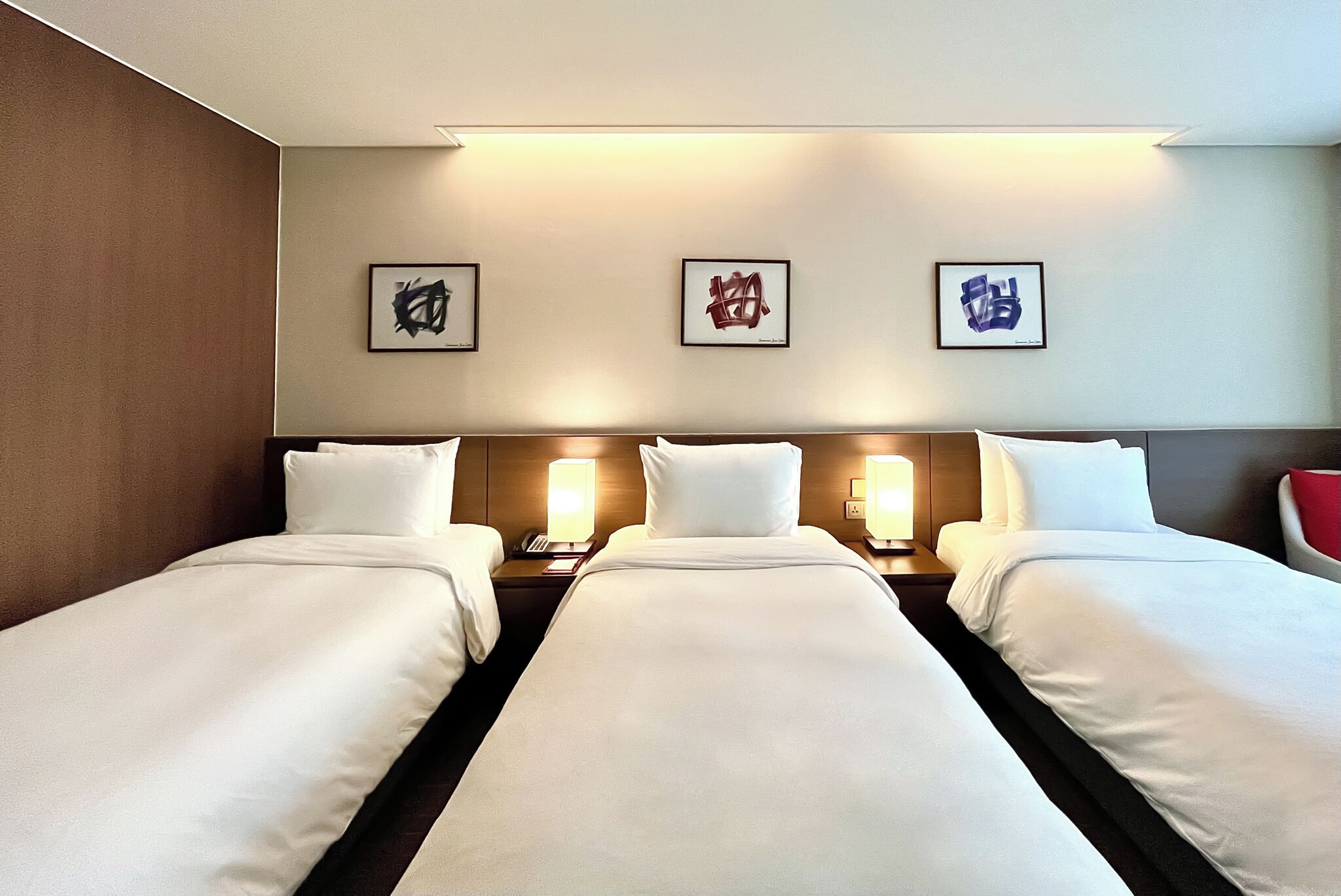 A hotel room with three single beds, each with a bedside table and lamp, and a wall with three framed artworks.