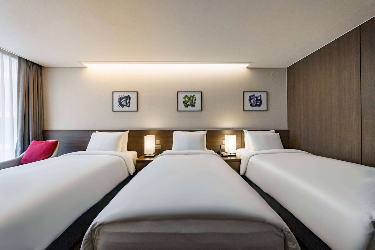 Habitación de hotel con tres camas, un ventanal con cortinas y tres cuadros enmarcados en la pared.