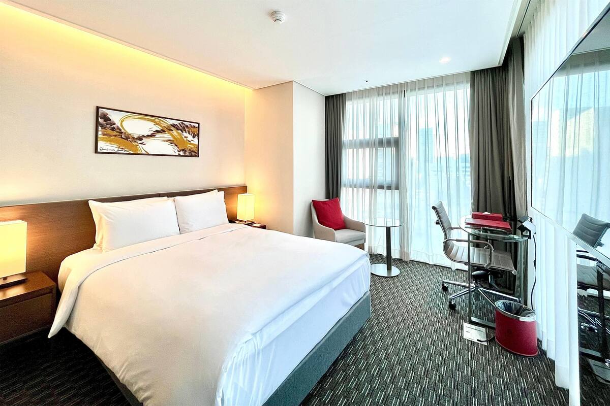 Habitación de hotel con una cama grande, un escritorio con silla, una mesita y una ventana con cortinas.