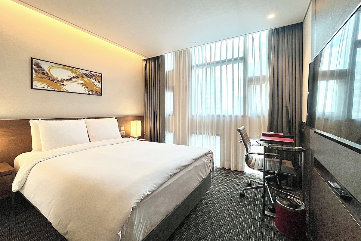 Habitación de hotel con una cama grande, un televisor de pantalla plana, un escritorio con una silla y una ventana con cortinas.