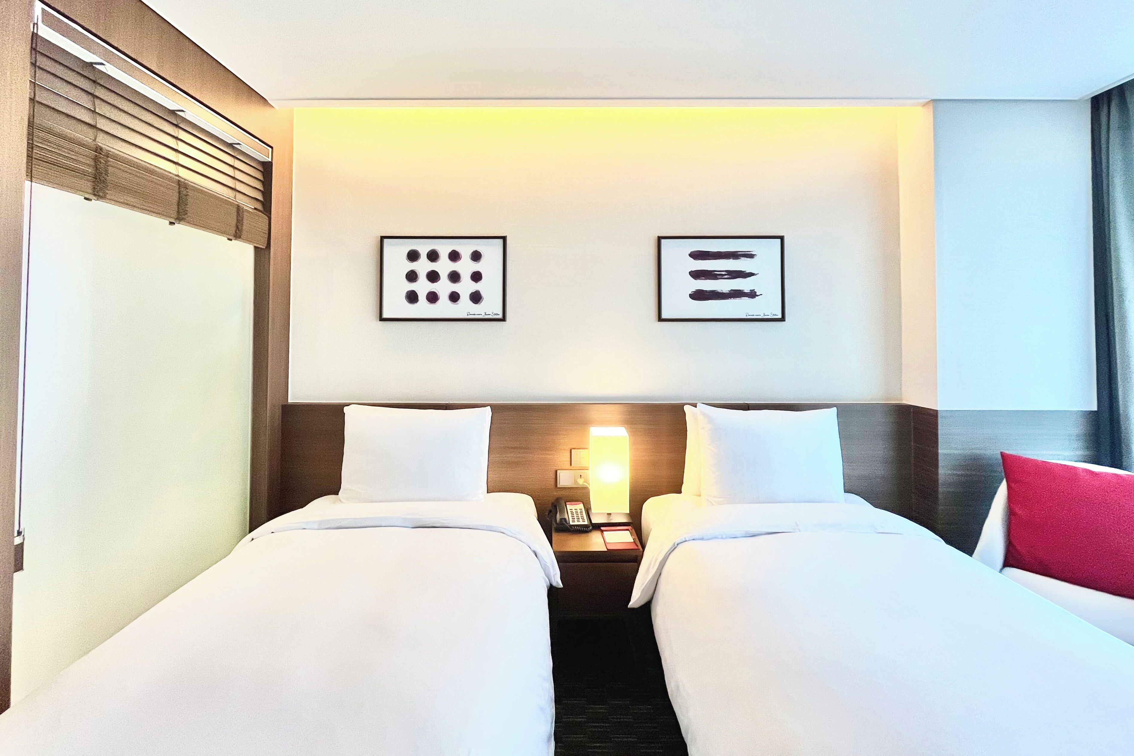 A hotel room with two single beds, a nightstand with a phone and lamp, and artwork on the wall.