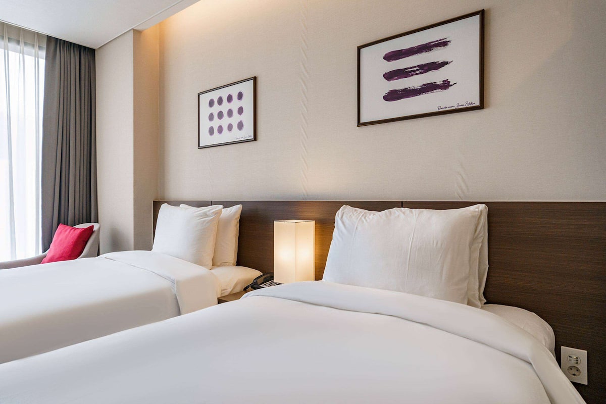 Habitación de hotel con dos camas, un cojín rojo, una lámpara de noche y dos cuadros enmarcados en la pared.