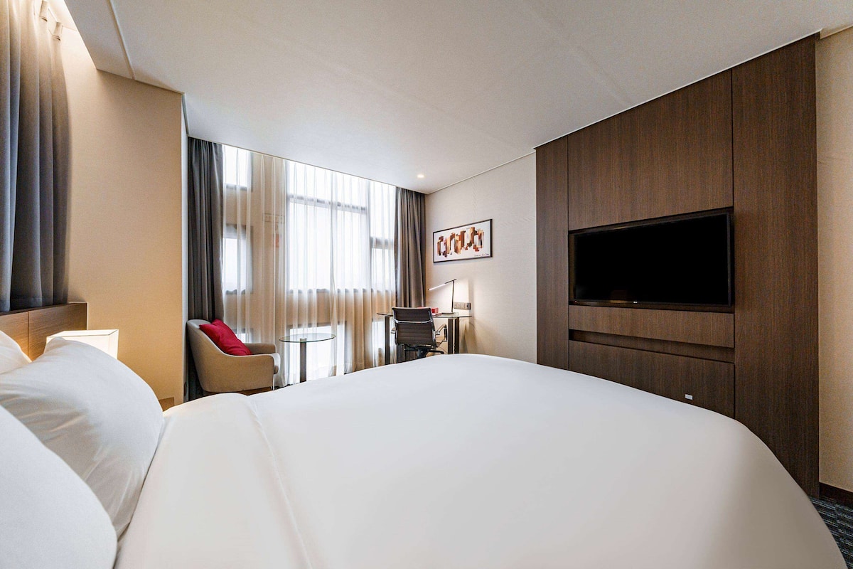 Una habitación de hotel con una cama grande, un televisor montado en la pared, un escritorio con una silla y una ventana con cortinas.