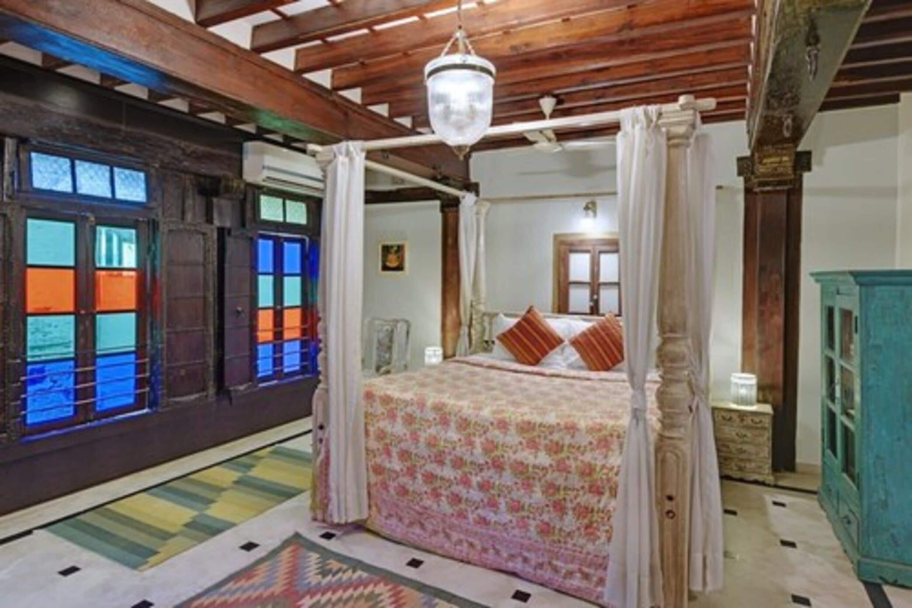 A four-poster bed with a floral bedspread, a wooden ceiling, and a view of a room with a green cabinet and a window with colorful panes.