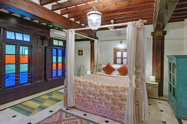Una cama con dosel adornada con una colcha floral, techo de madera y vista de una habitación con un aparador verde y una ventana con vidrios de colores.
