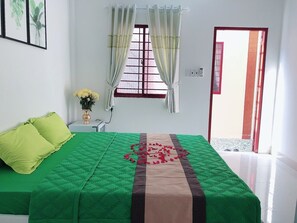 A bedroom with a green bedspread, a red and beige runner, a small bedside table with a vase of flowers, and a window with curtains. - Moonlight Home (Ho Chi Minh City)