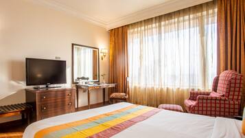 Habitación de hotel con cama, escritorio, televisor, silla y ventana con cortinas.