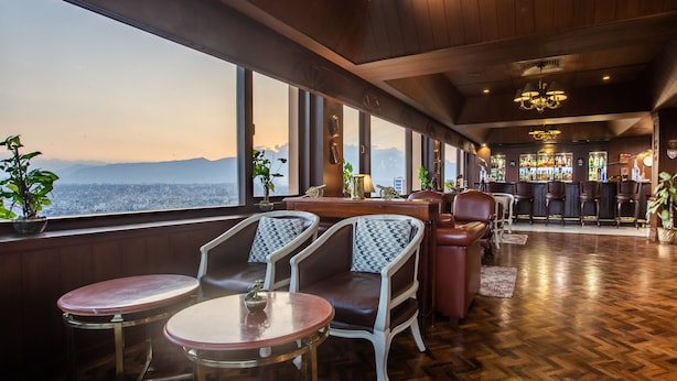 Un área de bar con suelo de madera, taburetes y vistas a la ciudad a través de amplias ventanas.