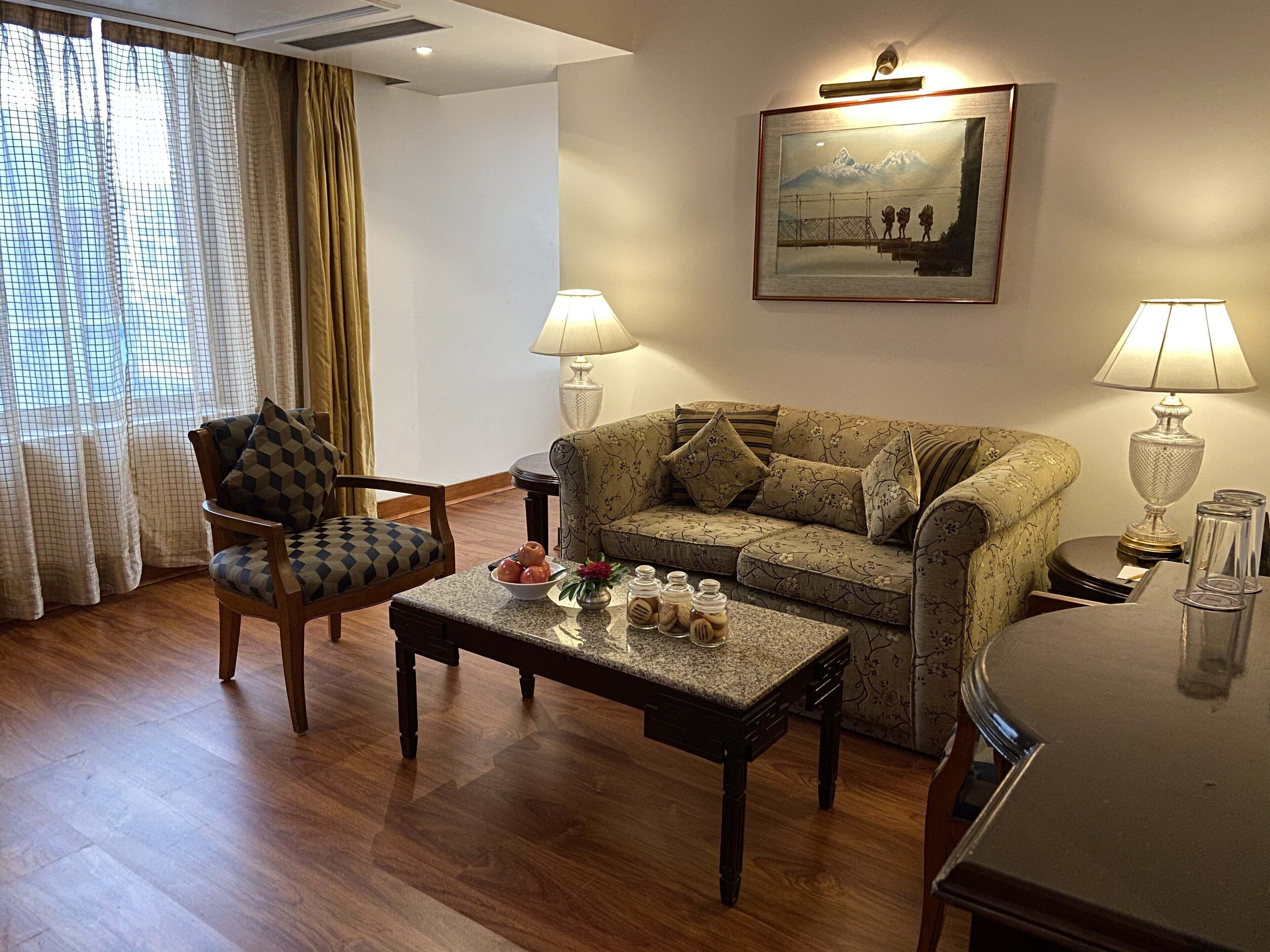 Een woonkamer met een houten vloer, een sofa met een patroon, een houten fauteuil, een bijzettafeltje en twee lampen.