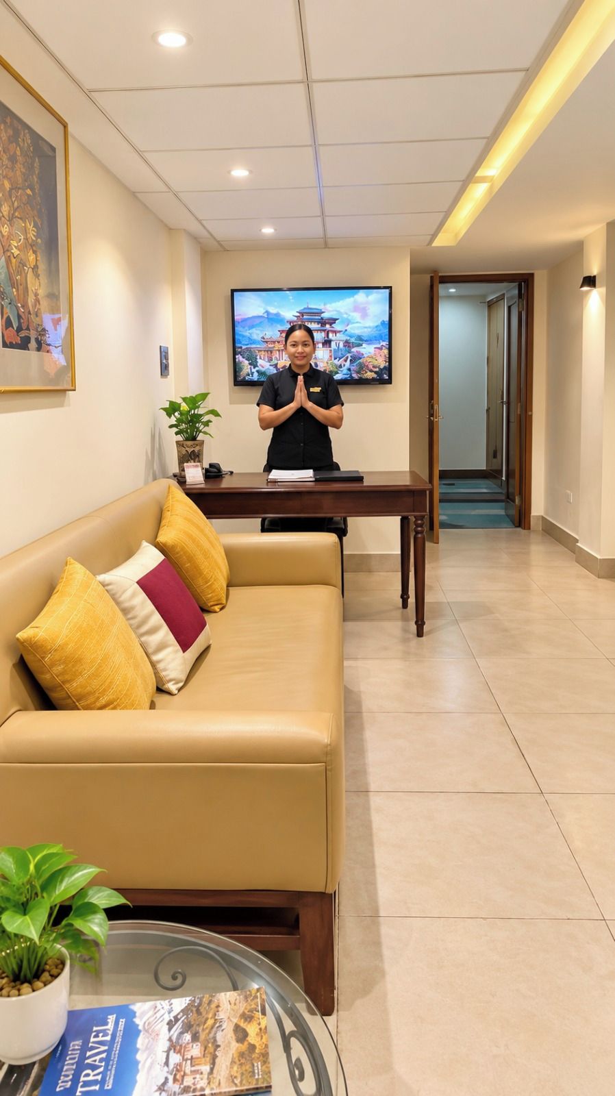 Ein Hotel-Rezeptionsbereich mit einer Rezeptionistin, einer Couch mit Kissen, einem Fernseher, der ein Landschaftsbild zeigt, und einer Zimmerpflanze.