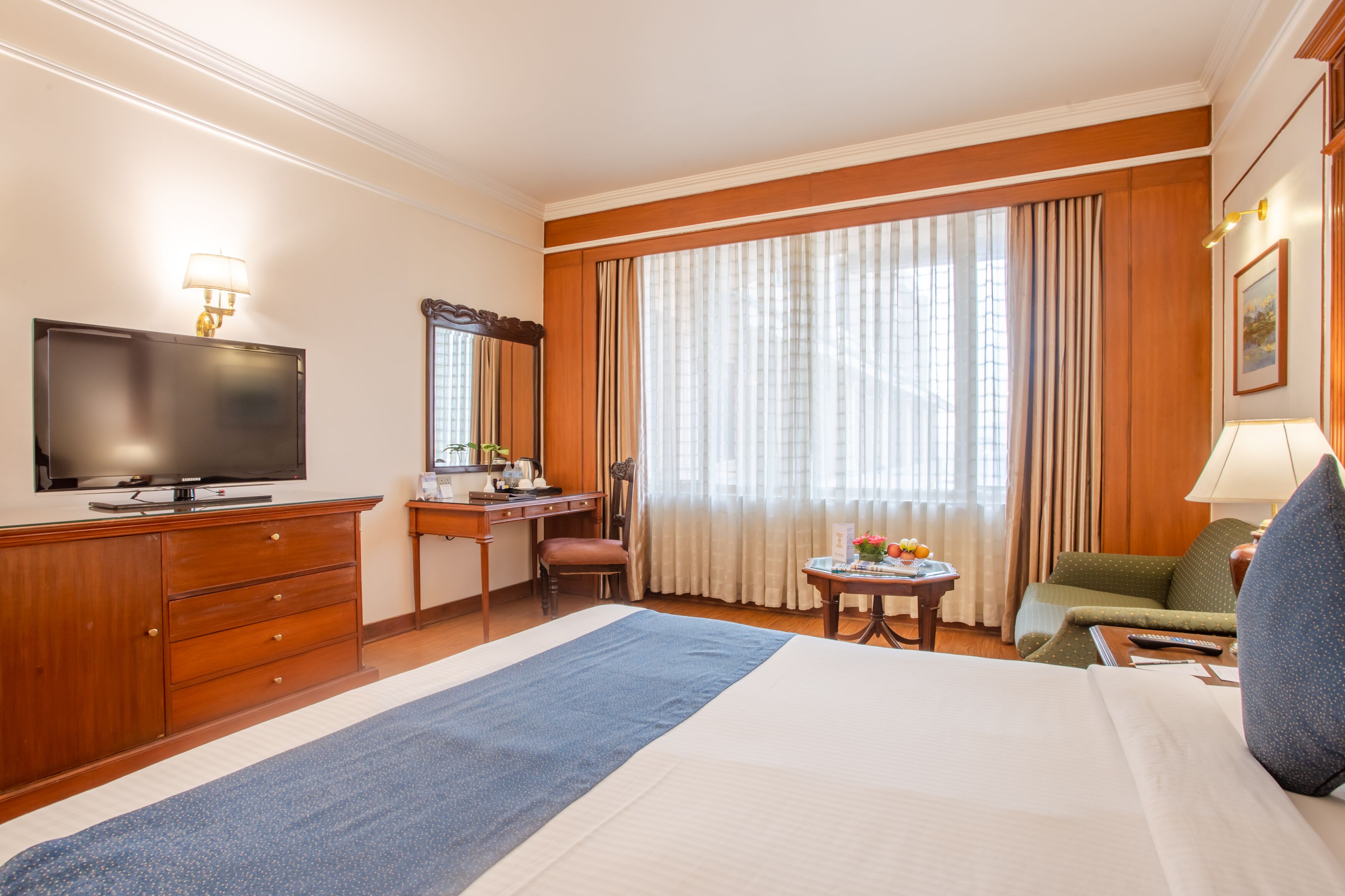 Habitació d'hotel amb un llit gran, una escriptori amb televisió, una cadira, una taula petita amb flors i una finestra amb cortines.