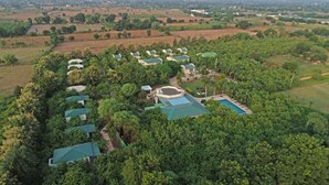 Um conjunto de edifícios com telhados verdes, rodeado por vegetação densa, com uma piscina e campo de tênis à vista.