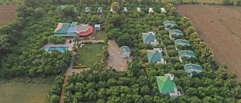 An aerial view of a resort with multiple buildings surrounded by dense greenery, a swimming pool, and a central area with a red-roofed structure.